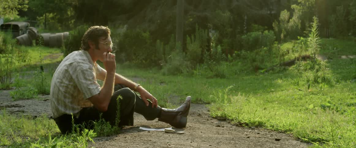 A man sits on a gravel rural pathway smoking a cigarette