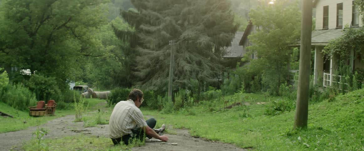 A man sits on a dirt road in front of his house and smokes a cigarette