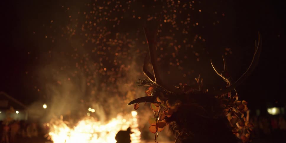 A person wearing a headdress of leaves and antlers watches a large bonfire along with a crowd at night