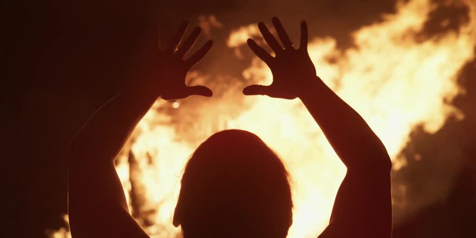 A person stands in front of a roaring bonfire with their arms and hands stretched into the air