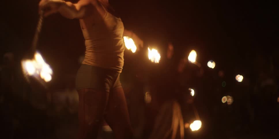 A man and woman dance outside at night with two batons lit on fire on both ends for a small crowd