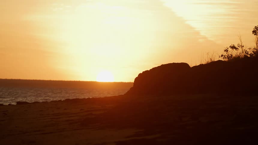 The sun sets over the sea and a hill is silhouetted against the red sky