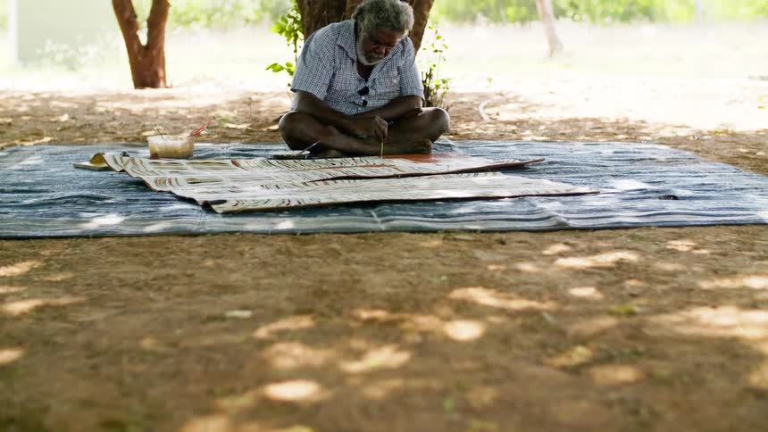 A man sitting down on a blanket outside painting with trees in the background