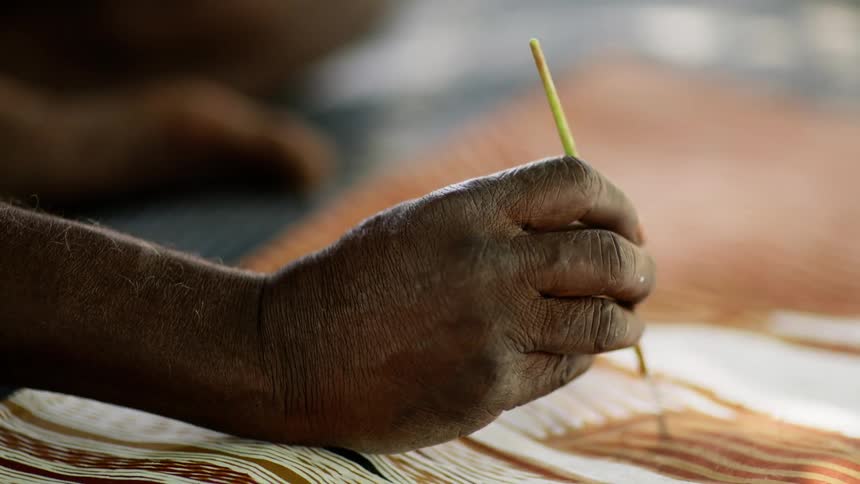 An elderly man sits on a blanket and uses a small paint brush to paint on a sheet of leather