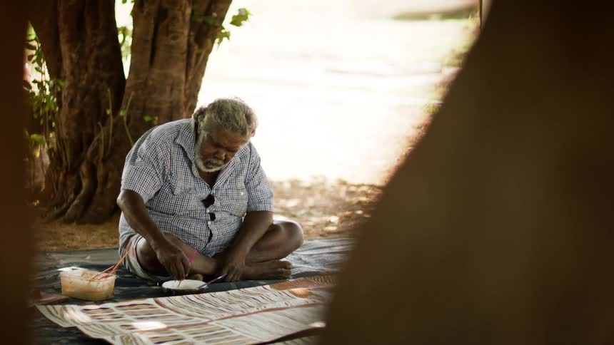 A man sits on the ground outside as he weaves an intricate rug