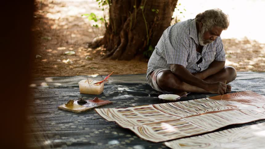 An elderly man sitting on a blanket outdoors paints on a leather hide
