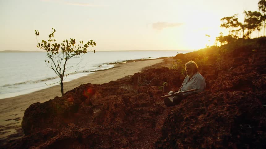 Man sitting cross legged on rocks above beach at sunset