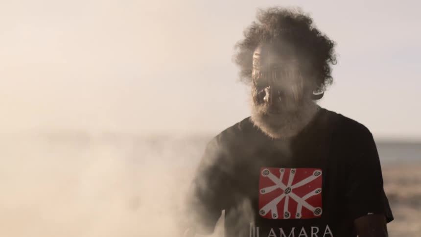 An elderly man wearing face paint sits in front of a smoking bonfire on the beach during the day
