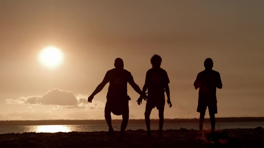 Three people stand around a camp fire at sunset