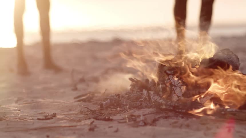Indigenous people stands around a fire on the beach next to the sea at dusk