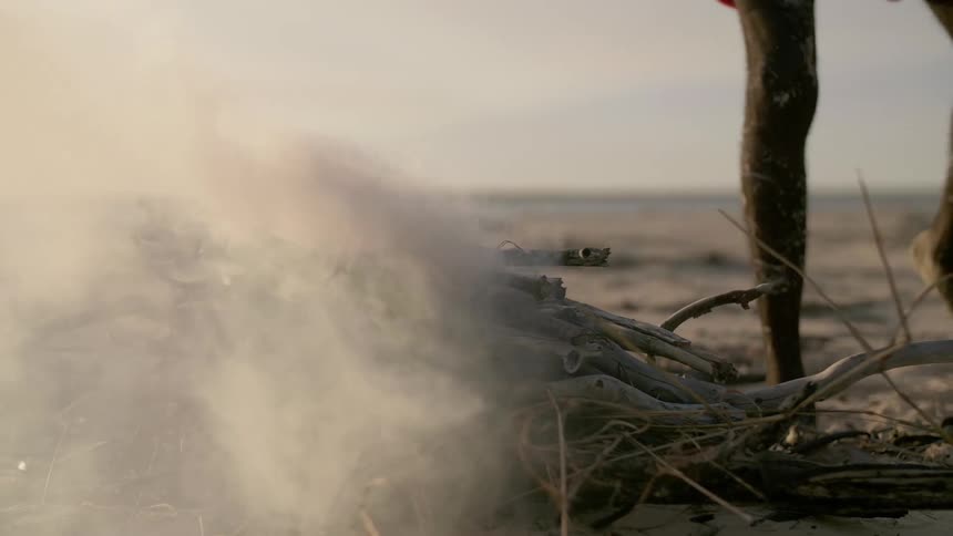 A man approaches and sits before a smoldering pile of twigs and branches on a beach