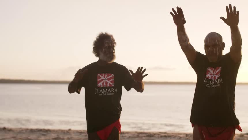 Two men wearing t shirts and face paint reach up as they move across beach with sun sitting on horizon