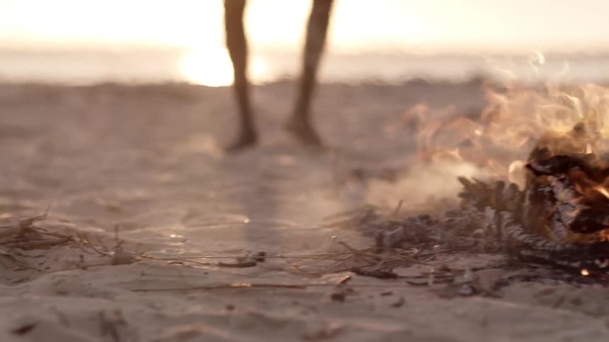 Group of men dancing around a camp fire on the sand of a beach next to the ocean at sunset