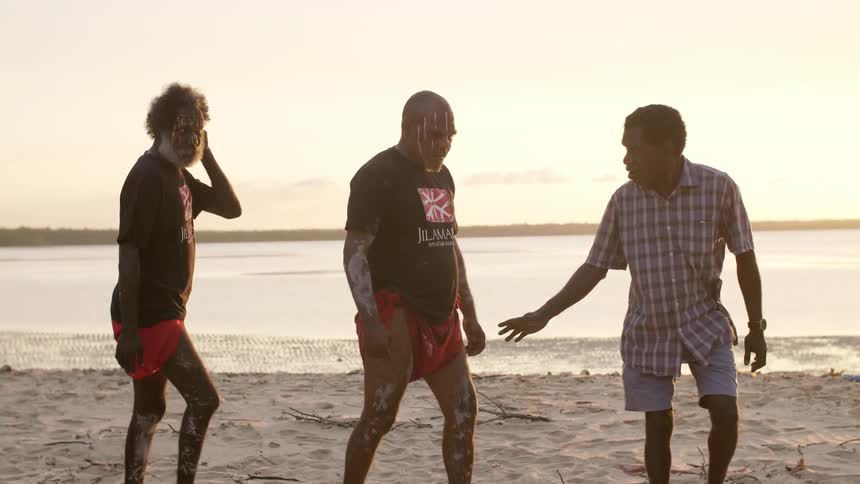 A group of elders wearing face paint walk in single file along a beach in the evening