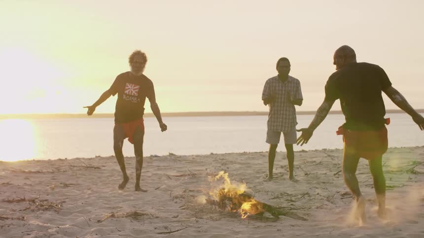 A group of australian aboriginal people dancing around a bonfire in the beach at sunset
