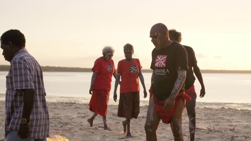 Tribe members walk around a fire on the beach
