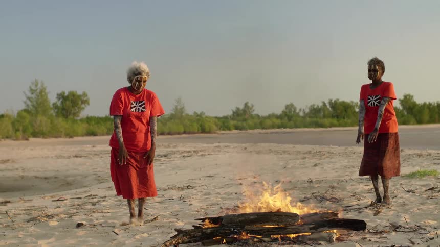 Two women wearing paint on their faces dance around fire on beach