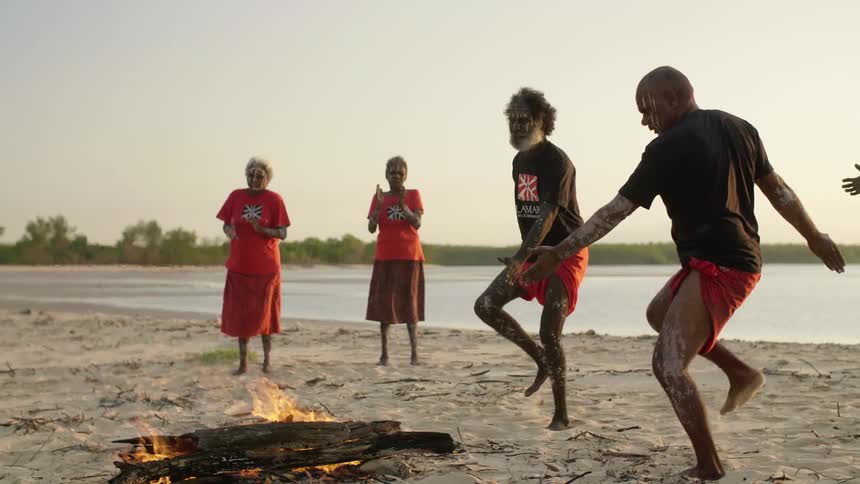 Two men do a tribal dance around a fire at three others stand and watch and clap