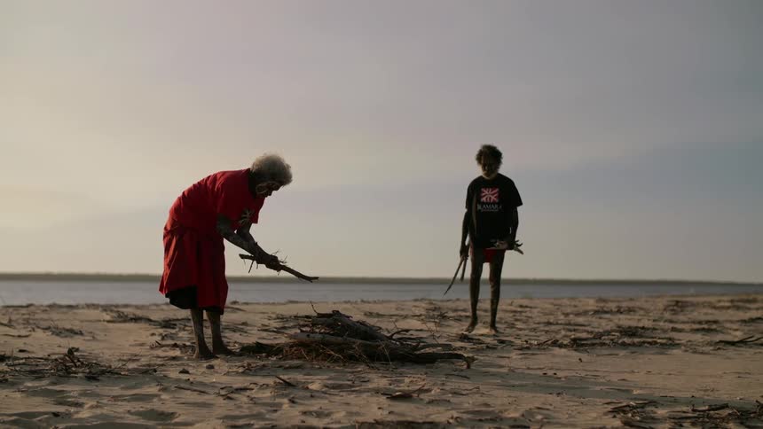 An elderly man and woman gather firewood and stack it on a beach during the day