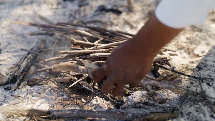 A man places sticks on a dying fire in the sand on a sunny day