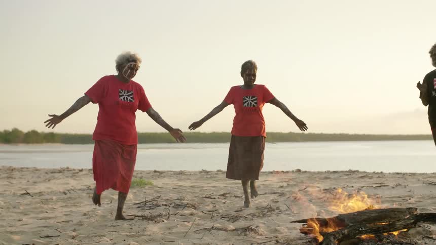 Two women with tribal paint on their faces dance around a fire on the beach as a man stands and claps