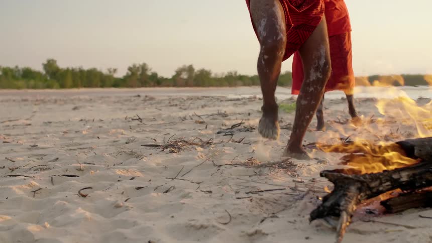 A group of indigenous people dances around a fire on the beach next to the sea at sunset