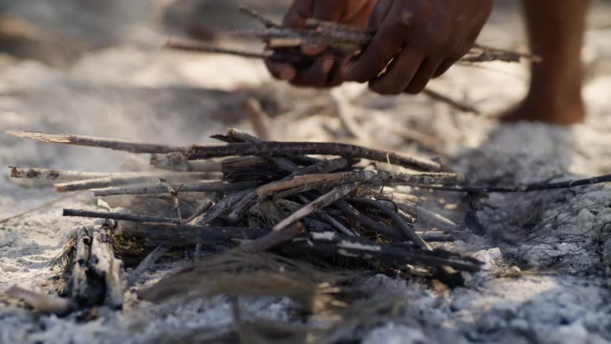 A man adds twigs to a pile to start a fire on the beach