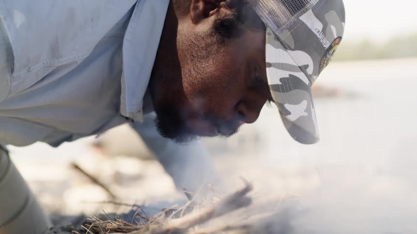 Man blows on small pile of smoking sticks to start fire