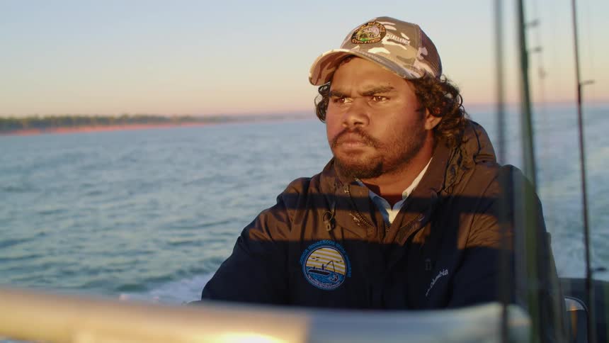 A man with a determined expression on his face drives a boat along the coast of a landmass at sunset