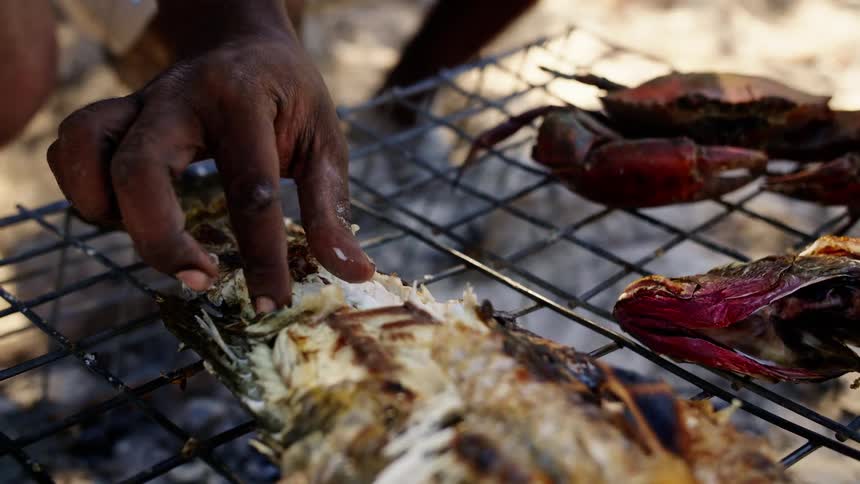 Two campers use their hands to eat a fish sitting on a wire basket with cooked crab