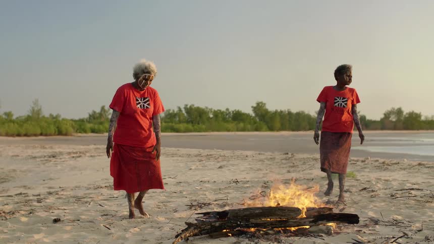 Two women with tribal paint dance around a fire on a beach as three men stand and watch