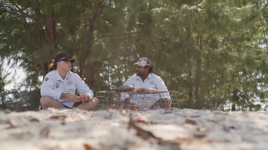 Two men sit on the beach and cook fish over an open flame