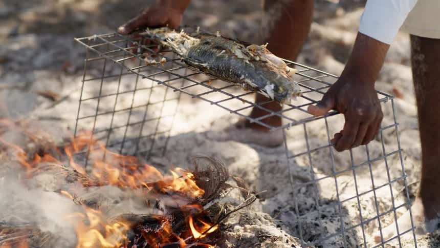 A man puts a fish over open flames outside to cook