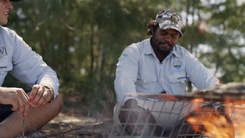 Two men sit cross legged on sand near food cooking on grill over campfire with trees at their backs