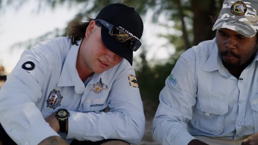 Two men sitting on the beach eat food cooked over a fire