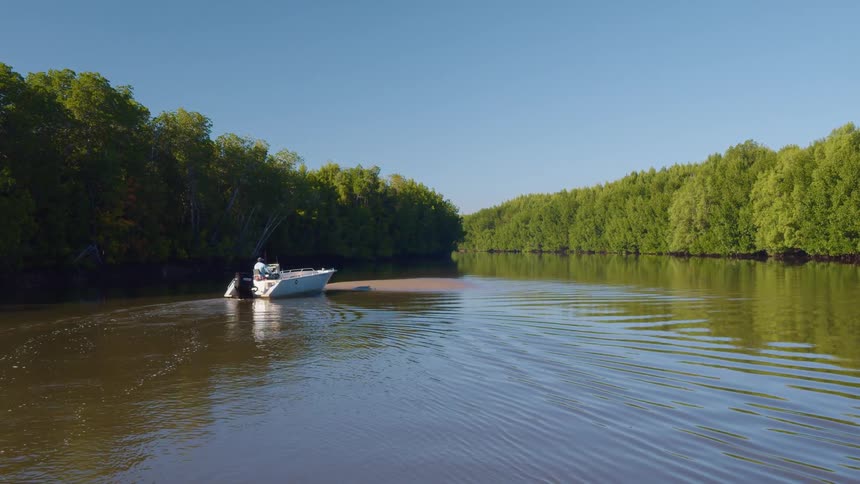 A man steers his boat as it slowly moves down a river surrounded by trees
