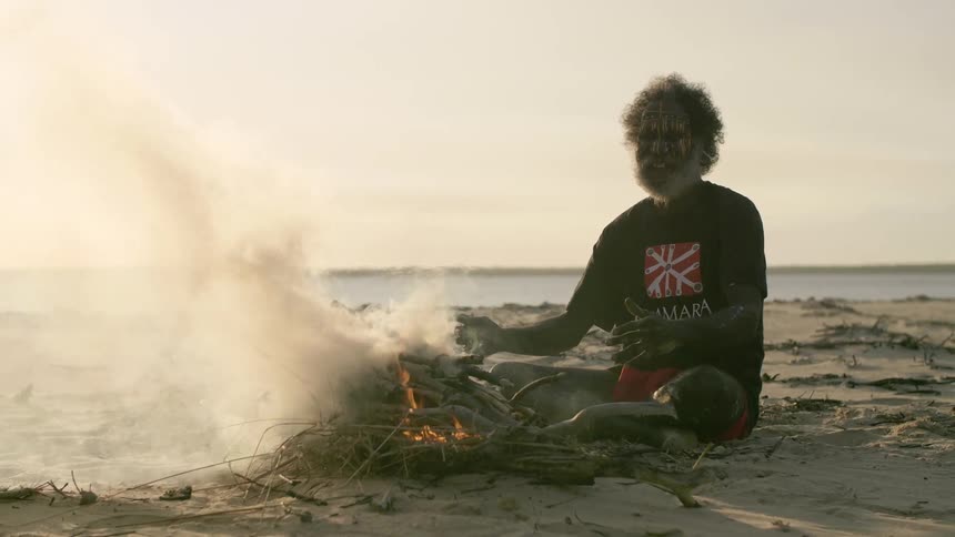 An elderly man with gray beard and face paint sits cross legged on the sand in front of a smoking fire
