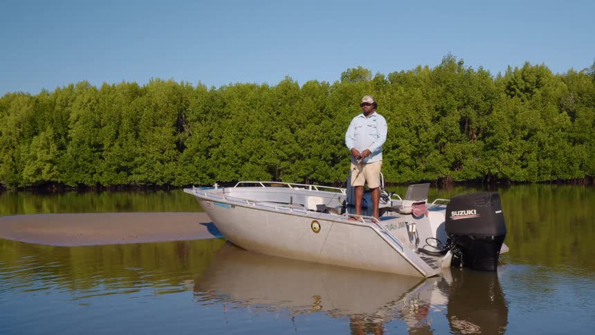 A man reels in his fishing line while standing on a boat in a murky body of water on a sunny day