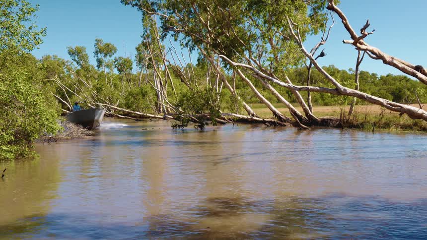A boater drives a boat along a muddy river through a grassland on a sunny day