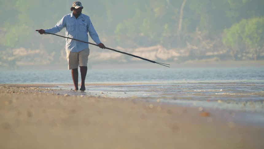 A man walks along the ocean edge carrying a large stick