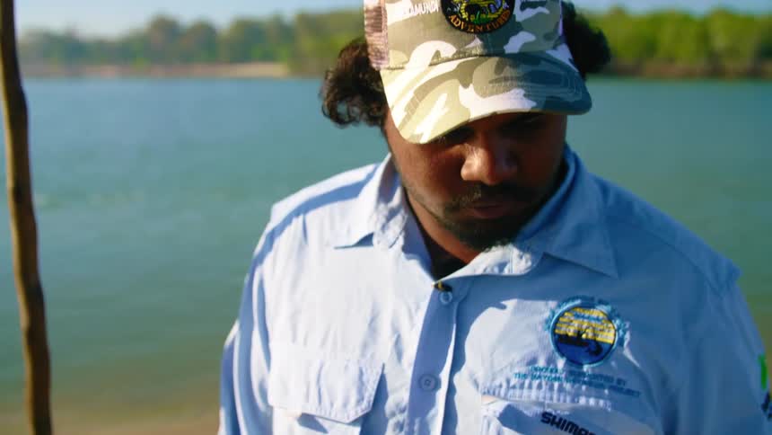 Man wearing baseball cap near ocean examines dead crab on sunny day