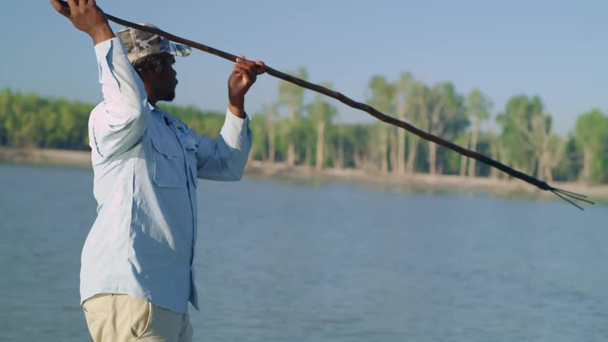 A man standing in the shallow waters of a lake throws a fishing spear into the water