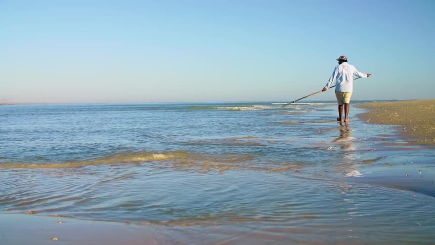 Man walks in water along beach carrying spear as water reflects his figure