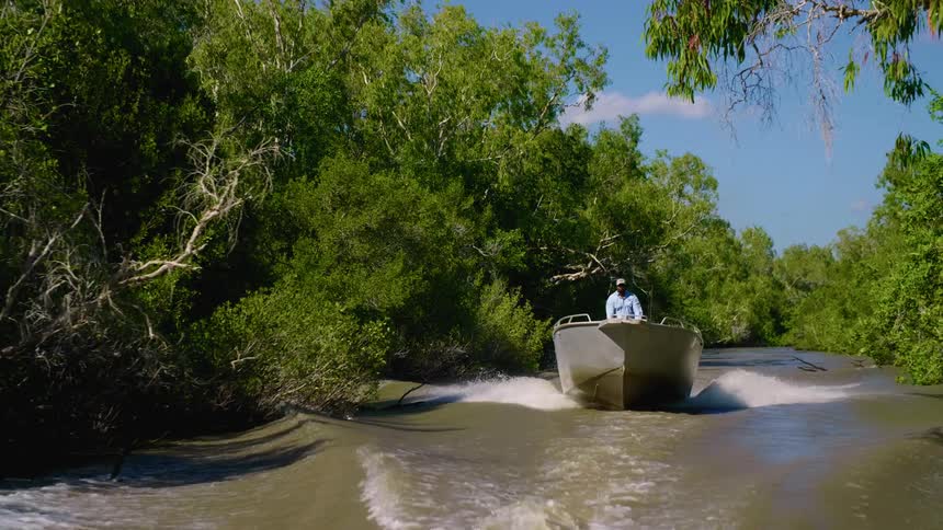 A man drives a motorboat through a murky marsh on a sunny day