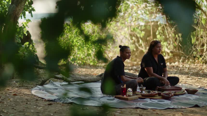 Two women sit on a blanket having a picnic with the ocean in the distance