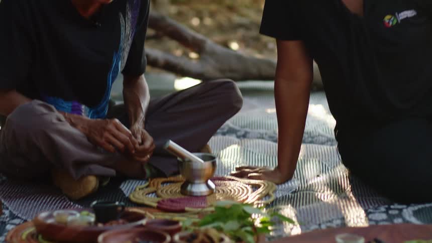 Two people sit on blanket near mortar and pestle discussing spices on tray