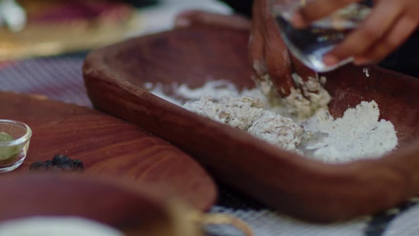 A person pours water into a wooden dish as they mix food in the wish with their other hand