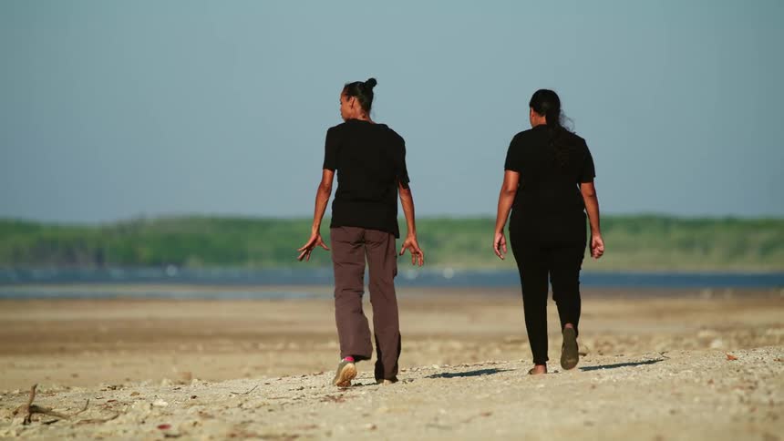 A pair of women chat while walking together along a beach under a clear daytime sky