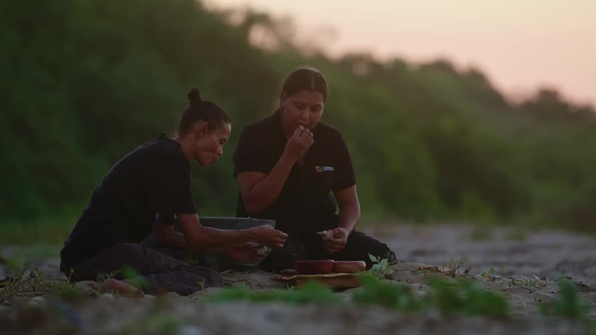 A pair of women sit on the ground outdoors while eating from a pair of bowls at sunset