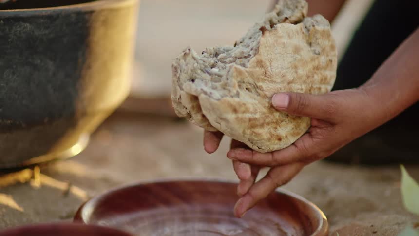 A woman kneeling down and breaking bread over a dish with trees and the sky in the background
